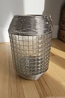 Full view of the glass lantern showing metal wire basketweave exterior and weighted bottom on wood floor with shadow cast by sunlight.