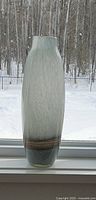 Tall slender modern glass vase on a window sill, showing semi-transparent grey textured glass with a weighted brown base.