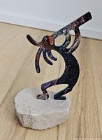 Side angle view showing multicolour Kokopelli figure in a bent posture playing flute, on top of natural sandstone base placed on wooden floor