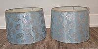 Pair of round pale bluish-grey lampshades with silver leaf pattern seen head-on against a white wall.
