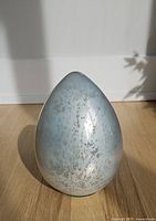 Front view of the pale blueish-grey mercury glass egg casting shadow on wooden floor against white wall.
