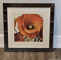 Front view of the framed print showing an orange poppy flower in a square mirrored dark brown frame against a light wall and wood floor.