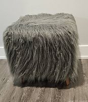 Shaggy faux fur foot stool cube with gray long faux fur and wooden legs on a wooden floor against a white wall.