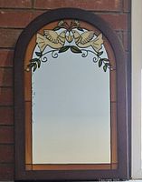 Front view of the arched stained glass mirror depicting two doves with olive branches, in a dark wooden frame against a brick wall.
