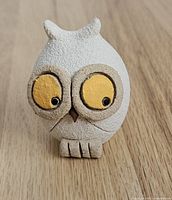 Front close-up of the cream colored owl figurine showing large yellow eyes and textured surface, standing on wooden floor.