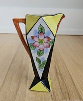 Mini jug front view on wood floor, showing hand-painted pink flower, geometric yellow and black shapes, and brown handle.