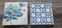 Two square lightweight wrapped canvas wall art pieces on wood surface. Left canvas shows a large blue flower and right canvas a blue and white geometric pattern.