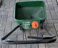 Front top-down view of green Scott Turf Builder Edgeguard Mini fertilizer spreader with orange adjustment dial and black handle, placed on stone tile floor.