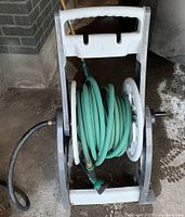 Photo showing white garden hose reel cart with black wheels; green garden hose coiled on reel with attached green spray nozzle at end.