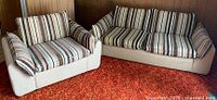 Vintage sofa and chair side by side showing striped cushions and beige base fabric upholstery on orange carpet background.