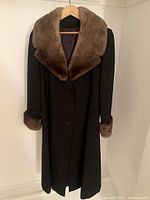 Front view of black wool coat with wide brown fur collar and fur trimmed cuffs, hanging on wooden hanger against white wall.