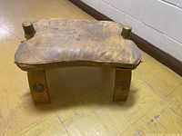View of foot stool showing distressed leather top, brass studs, brass medallions on wooden legs, and general wear pattern.