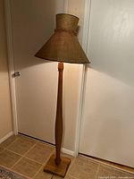 Full view of the floor lamp showing burlap shade with trim, wooden carved stand, and square base.