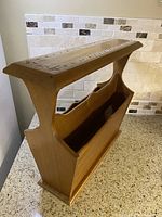 Side angle view of teak magazine rack showing curved profile and beadwork inlay on top.