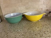 Two vintage Pyrex bowls, green and yellow, displayed side by side on countertop, showing their colors and designs clearly.