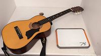 Half size acoustic guitar without strings placed on white surface, with attached black shoulder strap and whiteboard beside it.