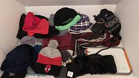 Photo showing assorted winter accessories including multiple tuques, a pair of gloves, and scarves in various colors and styles arranged on a white shelf.