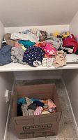 Photo showing a shelf with piles of infant and toddler clothing on top and a cardboard box filled with more clothes below.