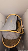 Opened Kidco Peapod travel bed showing grey fabric with orange-yellow trim, mesh sides, and padded floor
