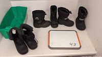 Four pairs of black kids winter boots from brands Sorel, Kamik and an unknown brand, sizes 8, 9, and 10 displayed on a white table with a green snow sled and small whiteboard marked 43.