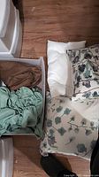 Photo shows a white pillow, two pillows with floral patterned covers, green fitted sheet and brown flat sheet on a wooden floor with plastic drawers nearby.