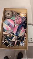 Top view of a cardboard box containing assorted ribbons on spools and multiple spools of thread in various colors and sizes.