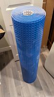 Photo of a blue rolled subfloor underlayment with circular bumps, standing vertically on a light gray wood floor near a white door and some cardboard boxes.