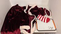 Photo showing a children's Christmas outfit including a burgundy satin skirt, matching top or cape with white faux fur trim, and red/white striped socks displayed on a white table.