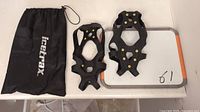 Pair of black IceTrax steel crampons with metal spikes beside a small white board on a table, along with the branded drawstring carrying bag.