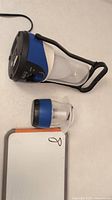 Two travel LED lanterns on white surface, large one with handle and black/blue casing, smaller one compact and cylindrical.