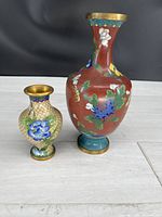 Two Chinese cloisonné enamel vases shown side by side on a light floor: one taller with red background and floral design, one smaller with beige crackled background and blue bird and flowers.