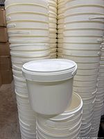 Stacks of white 1.3 gallon plastic buckets with lids, showing one bucket with lid on and another with handle up, illustrating design and quantity.