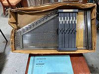 Autoharp placed in cardboard box with visible dust and a blue instruction manual below it, showing overall condition and packaging.