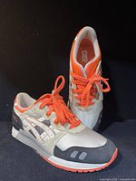 Pair of men's ASICS Gel Style III sneakers viewed from front and side angles on black background, showing cream and gray uppers with orange accents and laces.