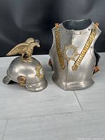 Photo shows both the metal cuirass and helmet side by side. The cuirass is approximately 13 inches tall with decorative gold straps and central emblem. The helmet has a gold eagle ornament on top and matching emblem on front.