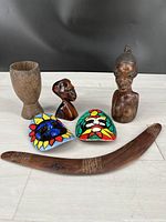 All six ethnographic souvenir items together on floor with black background: 2 carved busts, 1 carved cup, 2 painted masks, 1 boomerang.