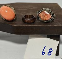 Three rings displayed on a wooden stand: peach/coral oval ring, dark wooden band, and orange stone ring with silver-tone setting.