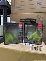 Two pairs of LG Tone Flex Bluetooth headsets in original boxes on cardboard box stack. Boxes show slight damage, items sealed.