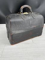 Front and side angled view of vintage black pebbled leather doctors bag, showing clasp and handle with visible wear and scuffs.