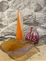 Photo showing three mid century art glass pieces against a stone wall background: orange/white sculpture, pink/white vase, amber colored bowl