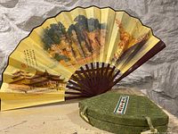 Yellow decorative folding fan with traditional Asian architecture and nature paintings, purple ribs visible. Green fabric-covered oval case next to fan.