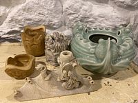 Photo showing five Asian vintage decorative items on a stone shelf: two small carved face-like pieces, one elaborate ceramic vessel shaped like a dragon, a small brown carved piece, and a long carved incense holder with dragons.