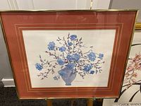 Framed watercolor painting showing an arrangement of blue flowers and red branches in a muted vase, mounted with pinkish mats and gold-tone frame.