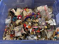 Wide view of mixed lot of vintage jewelry with many pieces jumbled inside a blue plastic container including pins, bracelets, and bead strands.