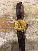 Front view of vintage Mickey Mouse watch showing gold-tone dial with Mickey Mouse on face and brown textured leather strap.