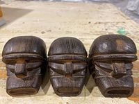 Photo of three mini wooden tiki masks carved with tribal features, shown side by side on a work table surface.