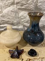 Photo showing a white alabaster lidded pot, blue-glazed pottery vase, and two small decorative glass pieces on a beige surface with stone wall background.