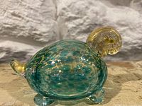 Side view of the Murano glass turtle showing turquoise and gold flecked body, golden head, and tail on clear glass feet.