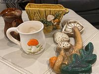 Group shot of the mushroom-themed ceramic items including mugs, wall plaque and decorative pieces on a white surface