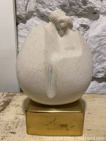 Front view of a light-colored textured stone sculpture of a stylized bird with carved feather details on the head, mounted on a polished brass square base, set against a stone wall background.